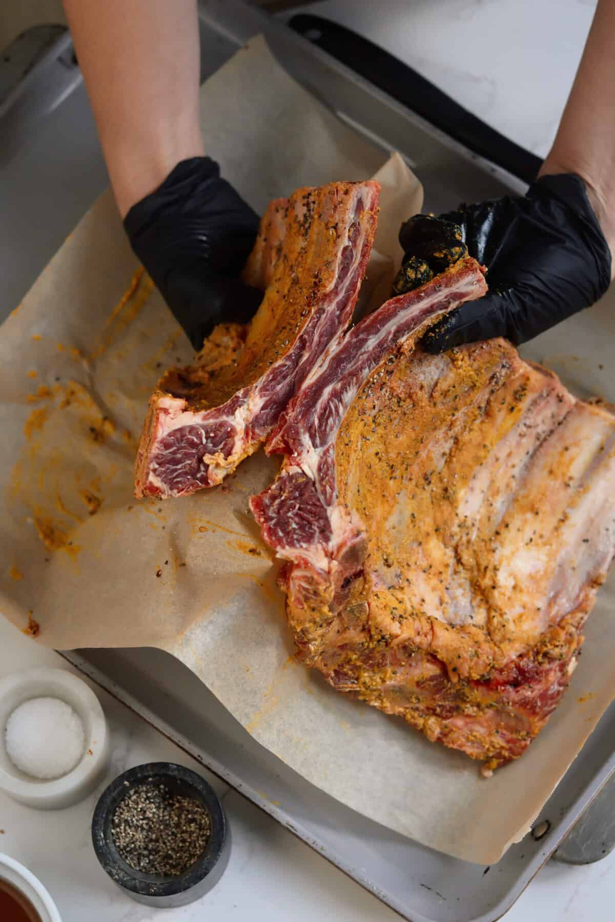 Overhead shot of raw seasoned baby back ribs being held and separated into two sections on a parchment-lined sheet pan so they can fit into the crockpot, with bowls of salt and black pepper nearby.