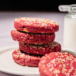 A stack of red cookies topped with colorful sprinkles sits on a white plate. A small glass jar, possibly of milk, is visible in the background on the right side.