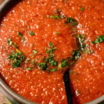 A close-up of a bowl of gazpacho soup, topped with chopped herbs and black pepper, with a spoon resting in the vibrant red soup.