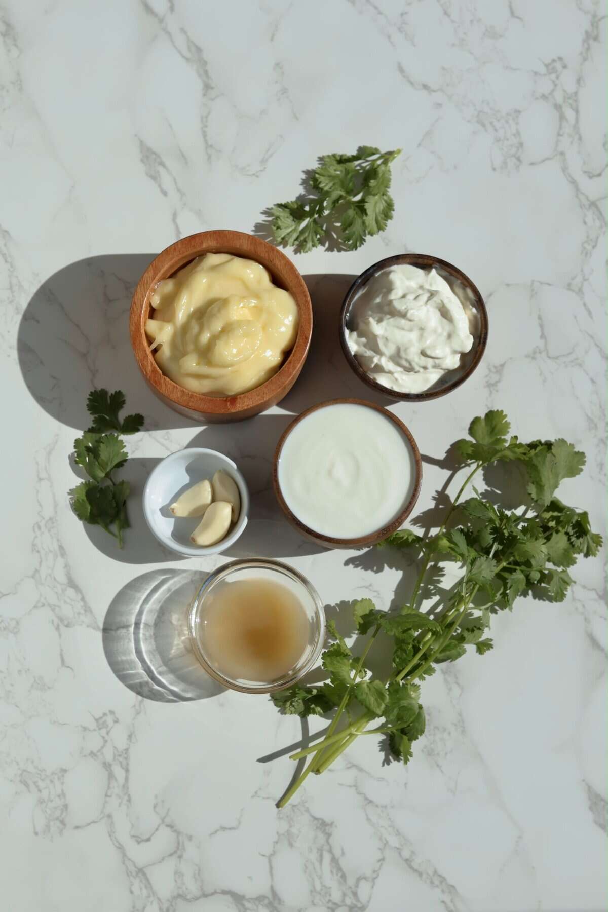 Overhead shot of garlic cilantro cream sauce ingredients on marble: mayonnaise, sour cream, milk, garlic cloves, lime juice, and fresh cilantro.