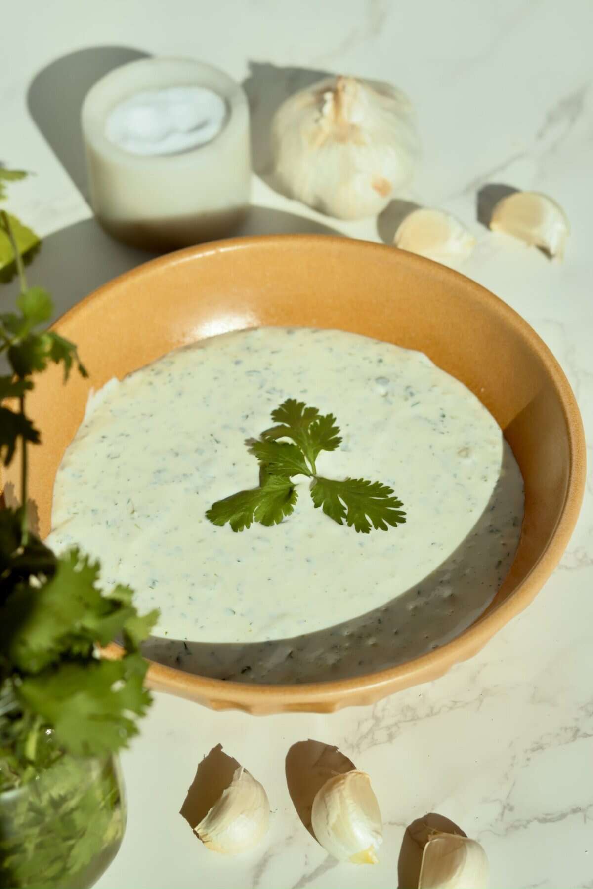 Close up of garlic cilantro cream sauce in mustard bowl garnished with fresh cilantro sprig on white marble surface.