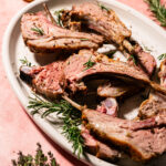 Sliced lamb chops garnished with rosemary on a white oval plate, with fresh herbs and a jar of olive oil in the background on a pink surface.
