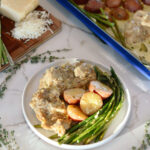 A plate with baked chicken, red potatoes, and asparagus. In the background, a baking dish with more chicken, potatoes, and asparagus, plus a cutting board with shredded cheese and asparagus.
