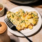 A slice of cheesy pasta casserole with herbs on a white plate next to a black fork. A pepper grinder and small bowl are nearby, with the casserole dish partially visible in the background.