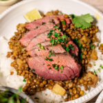 Sliced grilled steak served on a bed of white rice and lentils, garnished with chopped parsley and cilantro, with lime wedges on the side.