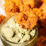 A fried shrimp is dipped in creamy green sauce in a small glass jar, with more fried shrimp pieces blurred in the background.
