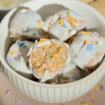 A bowl of white chocolate-coated treats topped with colorful sprinkles. One treat is bitten, showing a creamy, nutty filling inside. Some sprinkles are scattered on the surface around the bowl.