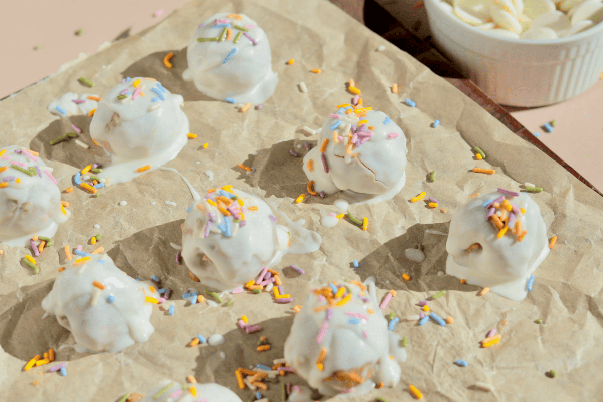 A glass bowl contains brown sugar pressed to one side, white liquid (likely milk or cream), and a splash of vanilla extract—perfect beginnings for no-bake dessert favorites like birthday cake truffles. Sprinkles decorate the surrounding surface.