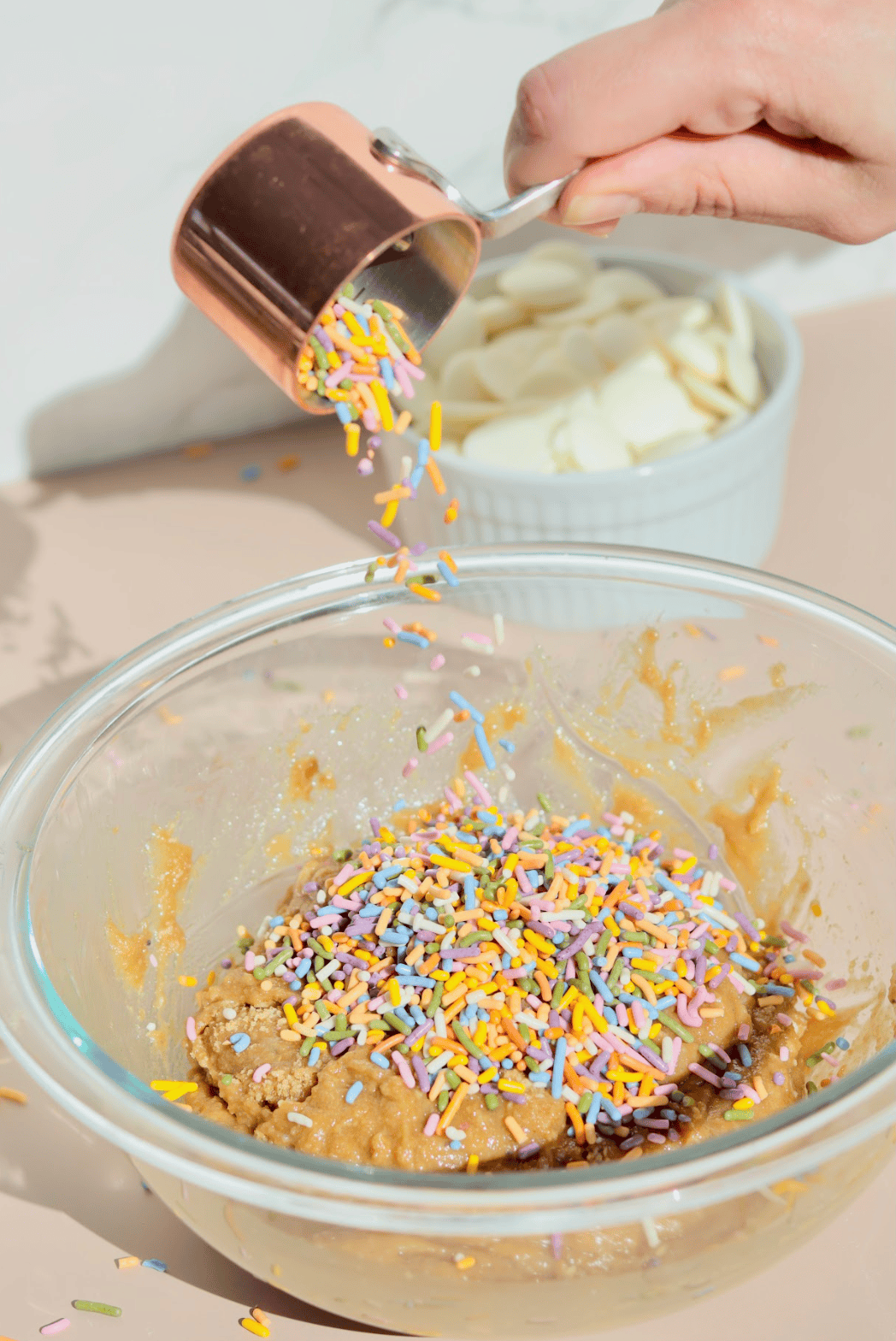 A glass bowl contains brown sugar pressed to one side, white liquid (likely milk or cream), and a splash of vanilla extract—perfect beginnings for no-bake dessert favorites like birthday cake truffles. Sprinkles decorate the surrounding surface.