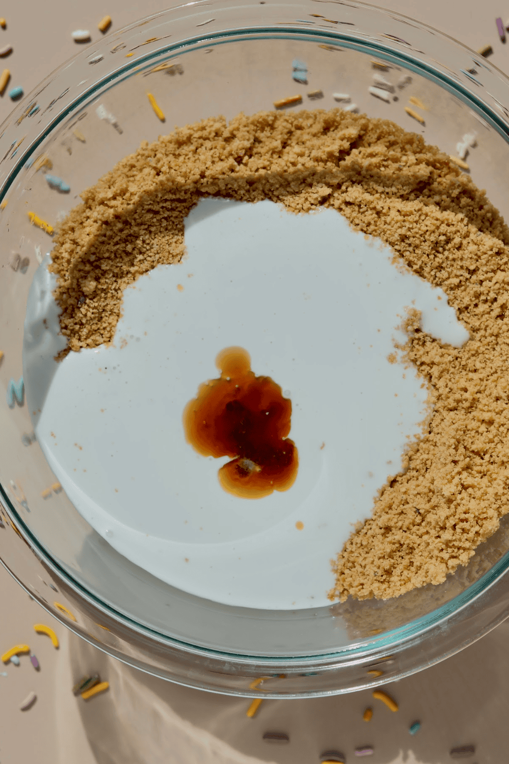 A glass bowl contains brown sugar pressed to one side, white liquid (likely milk or cream), and a splash of vanilla extract—perfect beginnings for no-bake dessert favorites like birthday cake truffles. Sprinkles decorate the surrounding surface.
