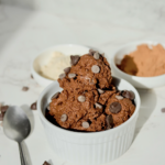 A white ramekin filled with scoops of chocolate cookie dough topped with chocolate chips. Two small bowls and a spoon sit nearby on a white surface.