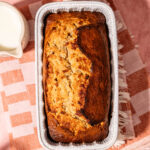 A loaf of banana bread in a white ceramic pan sits on a pink and white checkered towel, with a small pitcher of milk nearby on a pink surface.
