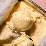 A close-up of a metal container filled with pale yellow mango ice cream, with one scoop resting on top and another scoop in a metal scooper.