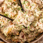 A bowl of creamy potato salad with chopped herbs, green onions, and two black serving utensils. The salad features chunks of red-skinned potatoes mixed with mayonnaise and garnished with fresh dill.