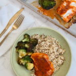 A green plate with seasoned white rice, roasted broccoli, and a piece of fish topped with red sauce—an inviting Asian Cod Recipe. In the background, a baking tray holds more broccoli and fish. A gold fork and knife rest on a napkin nearby.