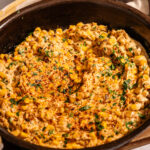 A skillet filled with creamy, seasoned Mexican street corn topped with chopped cilantro and spices, with a bowl of fresh cilantro and lime wedges in the background.