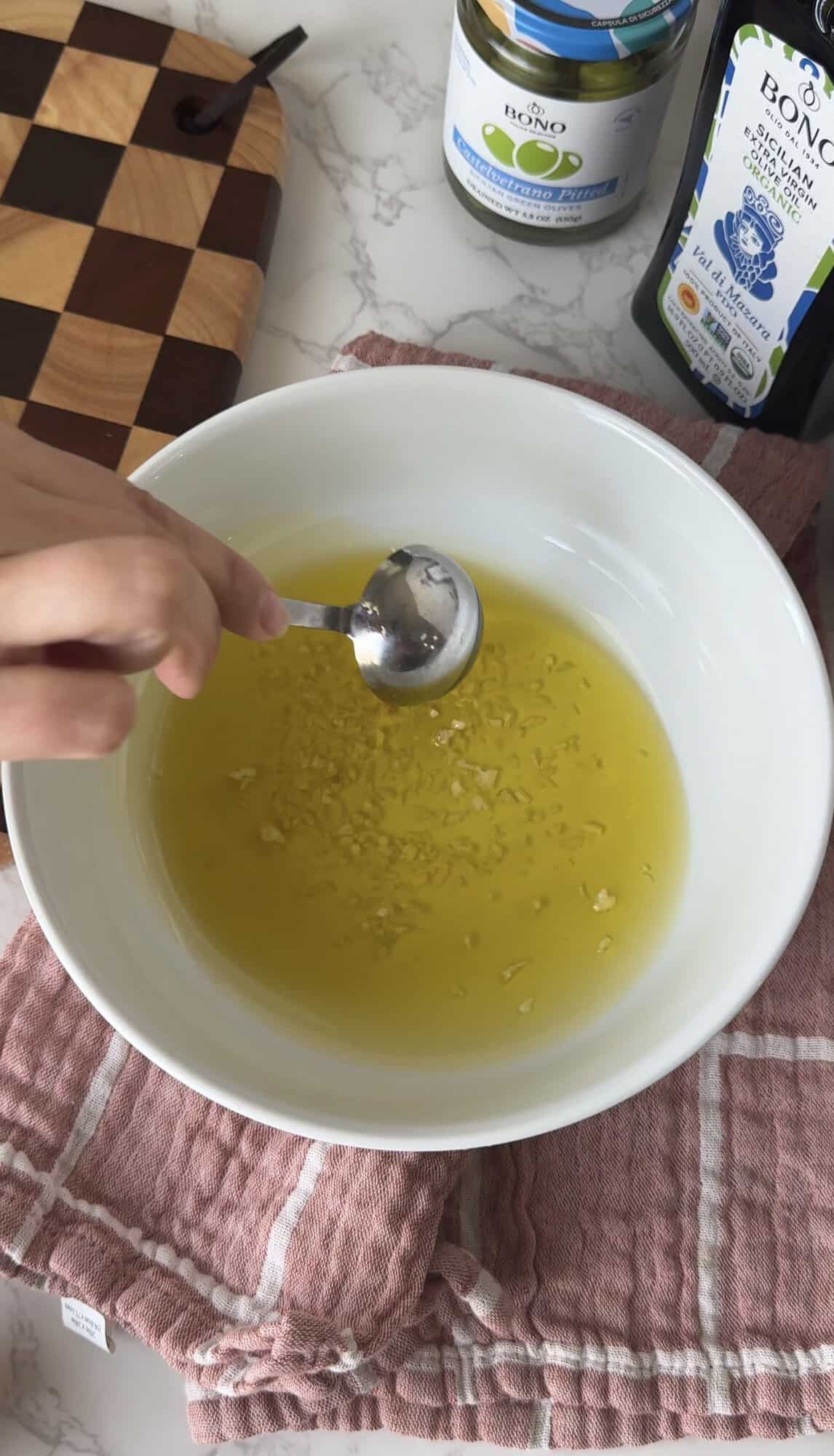 Stirring minced garlic into Bono Sicilian extra virgin olive oil in a white bowl to build the base for Italian herb dipping oil