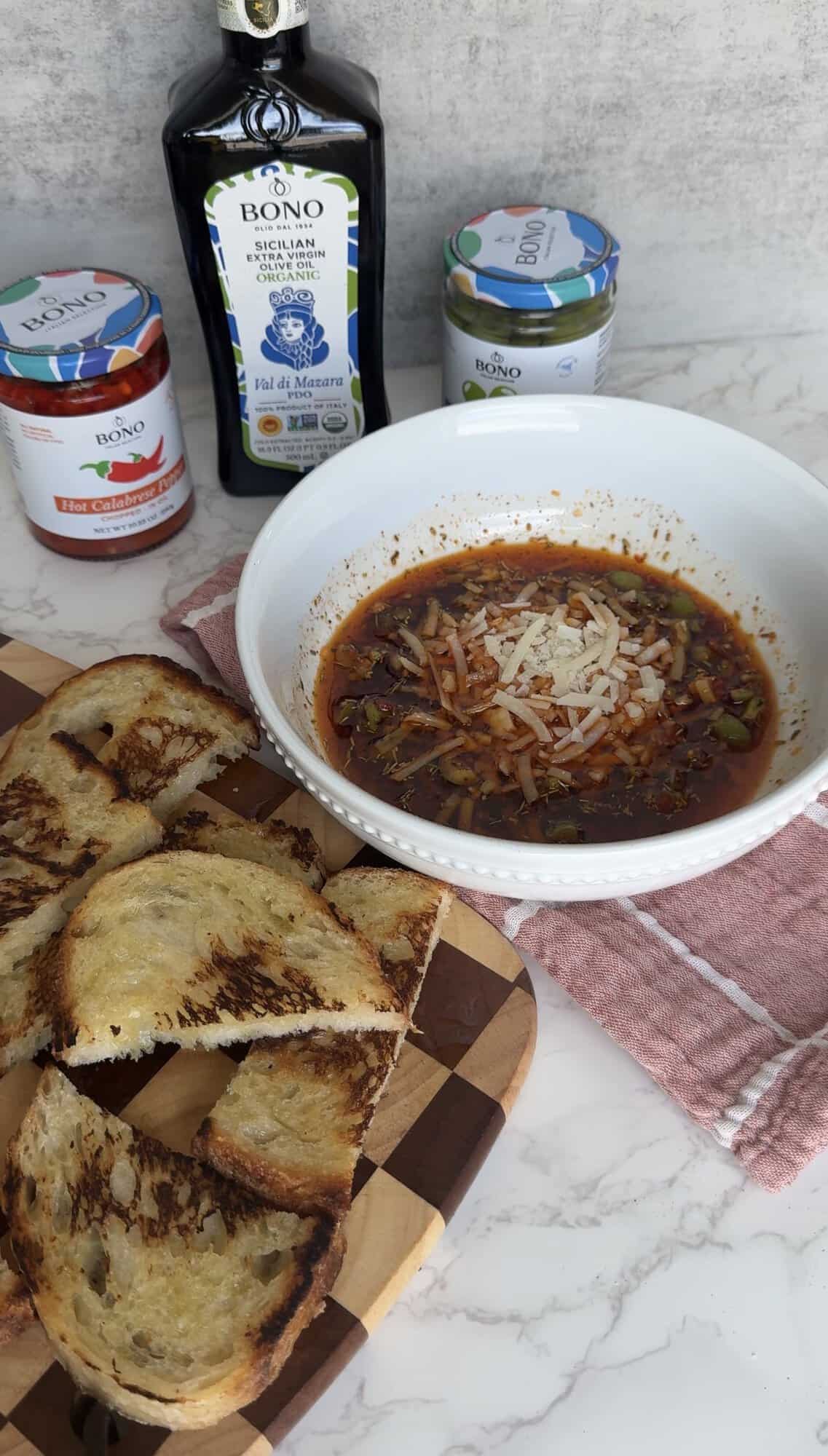 Italian bread dipping oil with shredded Parmesan served in a white bowl alongside toasted bread slices and Bono olive oil and Calabrese pepper jars