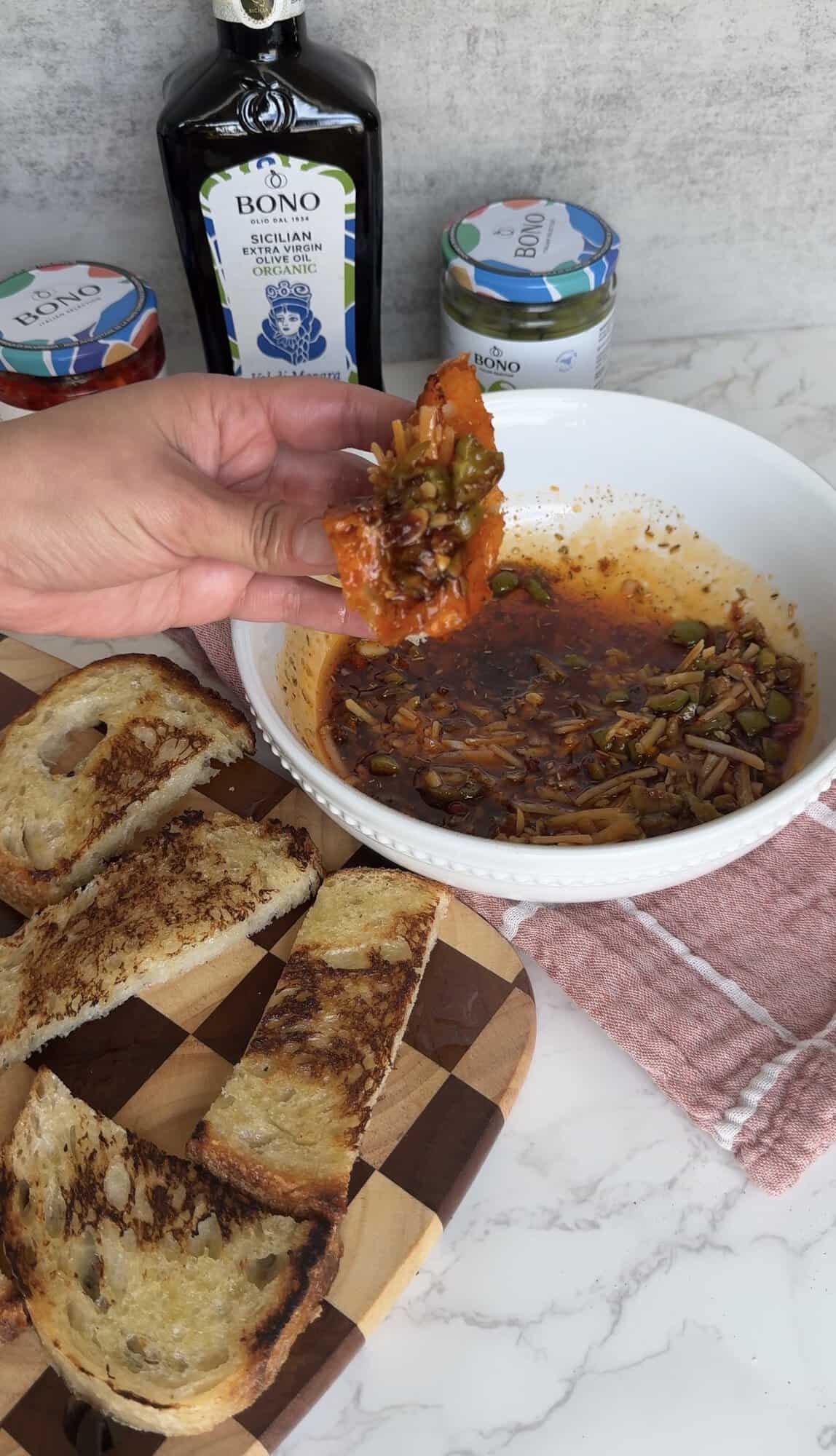 Hand holding a piece of toasted bread loaded with Italian herb dipping oil, olives, and balsamic