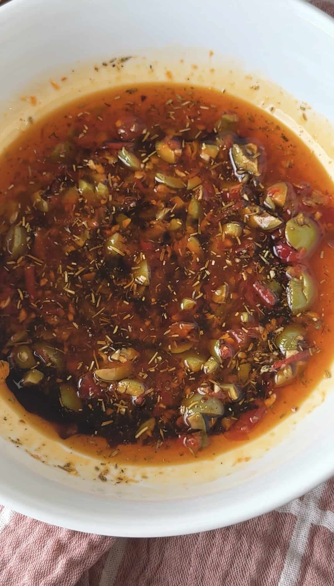 Loaded Italian herb bread dipping oil with Castelvetrano olives, Calabrese pepper, and balsamic vinegar in a white bowl