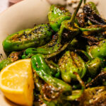 Charred green peppers in a bowl, reminiscent of blistered shishito peppers, garnished with black sesame seeds and served with a lemon wedge. A small bowl of black sesame seeds is in the background.