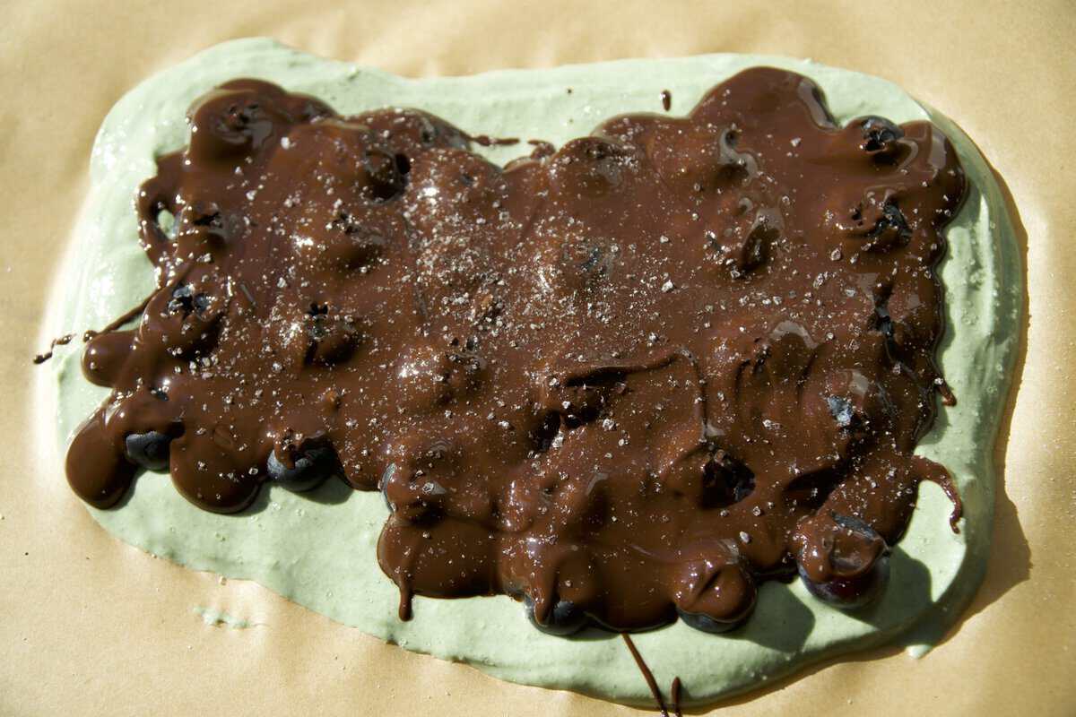 Melted dark chocolate spread over green Greek yogurt bark with blueberries and flaky salt on parchment paper