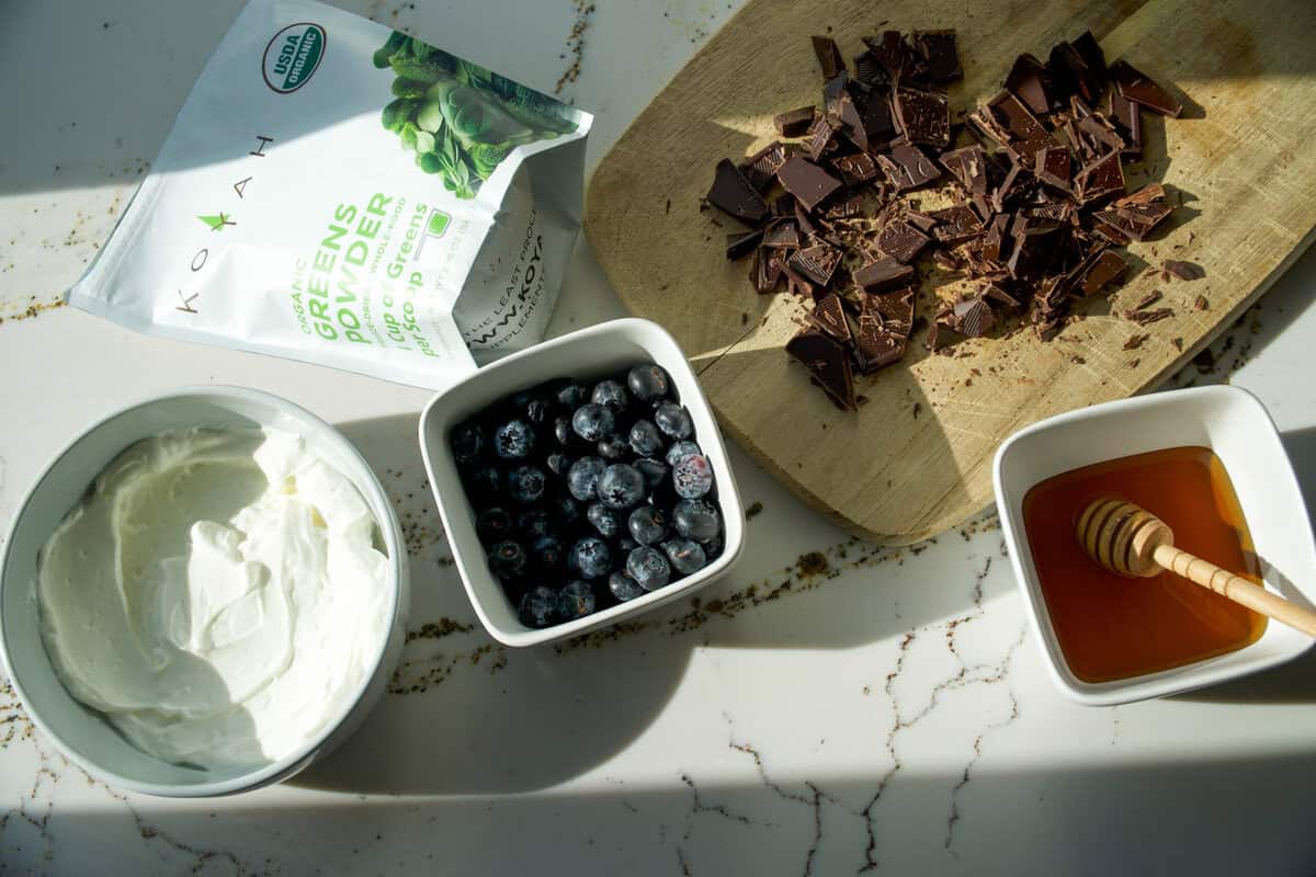 Greek yogurt bark ingredients flat lay with Greek yogurt, fresh blueberries, honey, chopped dark chocolate, and KOYAH greens powder.