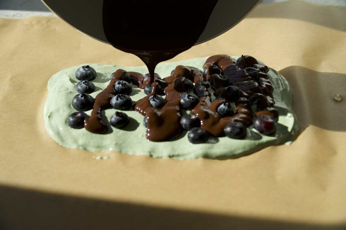 Pouring melted dark chocolate over fresh blueberries on green Greek yogurt bark