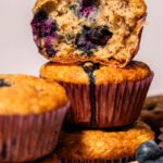A close-up of stacked blueberry muffins in red liners, with one muffin on top cut in half to show the moist, berry-filled interior. Fresh blueberries are visible on the side.
