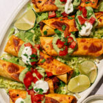 A platter of crispy Beef Taquitos with Avocado Sauce, topped with sour cream, diced tomatoes, and cilantro, served on a bed of shredded lettuce with lime wedges on the side.