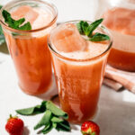Two glasses of strawberry lemonade with ice cubes and mint sprigs on top. Fresh strawberries and mint leaves are scattered nearby. A pitcher with more lemonade is in the background.