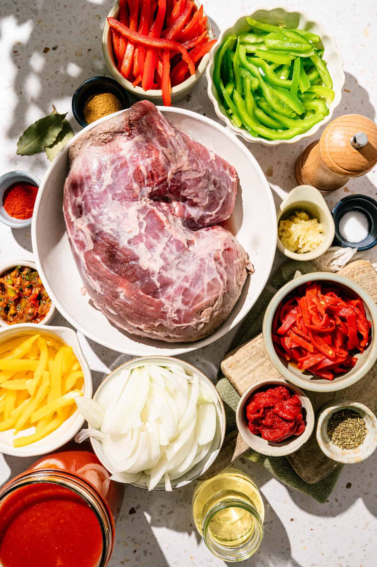 A bowl of raw meat surrounded by sliced red, green, and yellow peppers, onions, garlic, tomato paste, spices, wine, and other ingredients on a light-colored countertop.