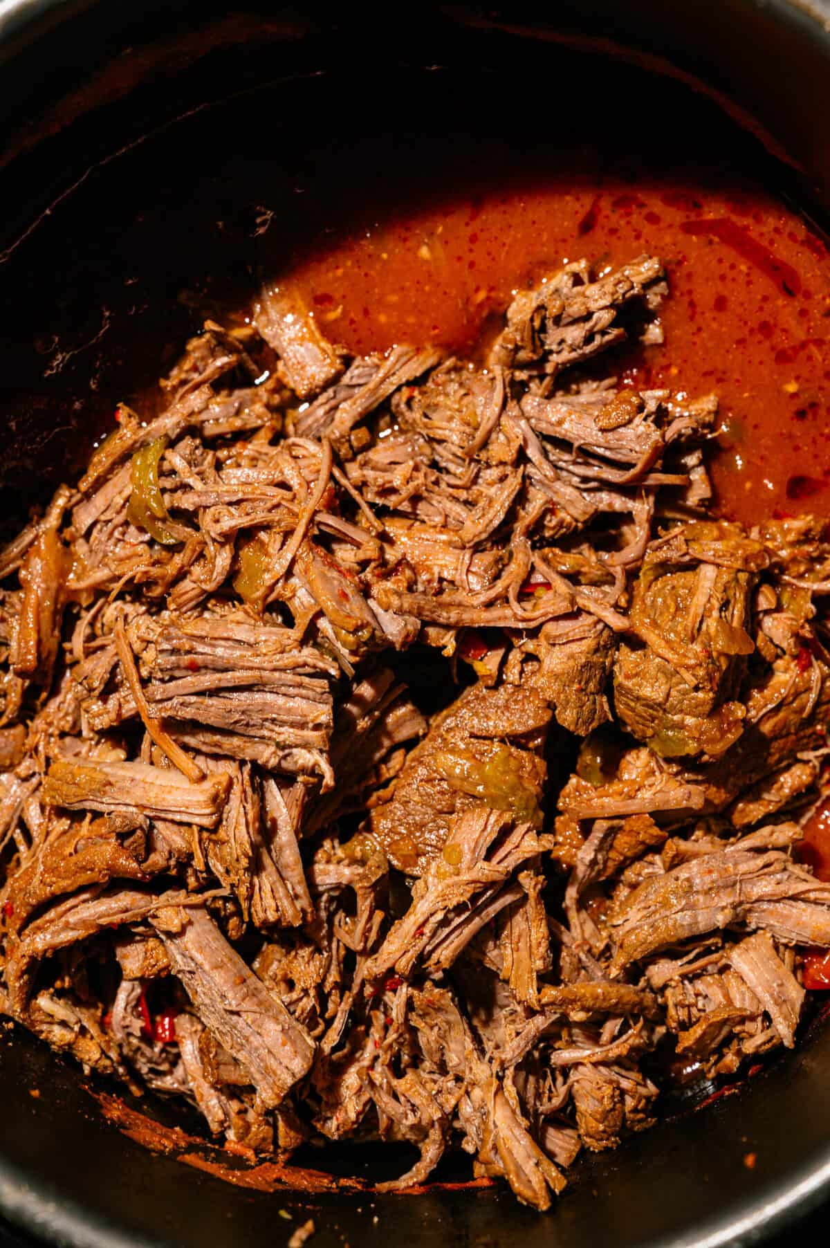Shredded beef in a rich, reddish-brown sauce with visible pieces of green bell pepper and onions, served in a dark pot.