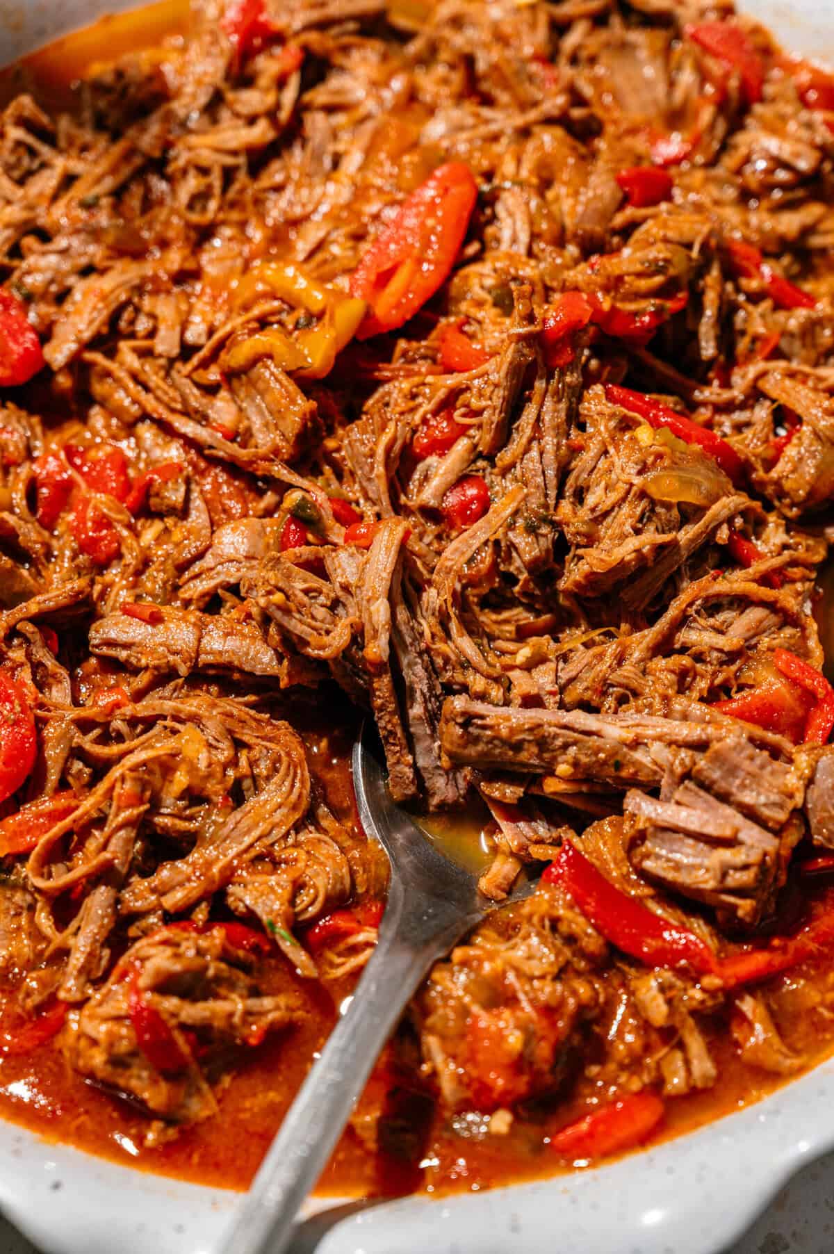 Shredded beef in a rich, red sauce with sliced red peppers, served in a white dish with a metal spoon.