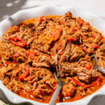 A white dish filled with shredded beef in a red sauce with sliced red peppers, served with a spoon.