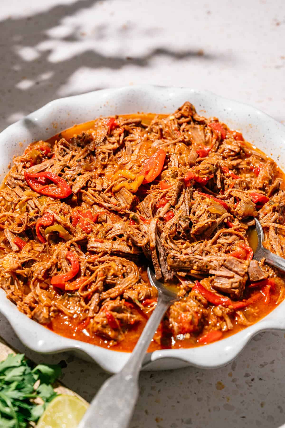 A white dish filled with shredded beef in a red sauce with sliced red peppers, served with a spoon.