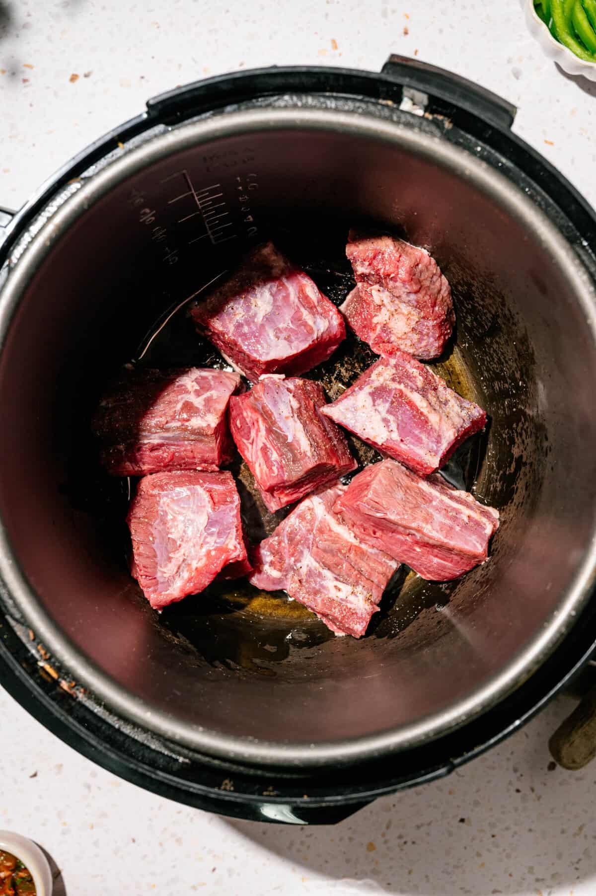 Chunks of raw beef short ribs are placed at the bottom of a black pressure cooker or Instant Pot, ready to be cooked.