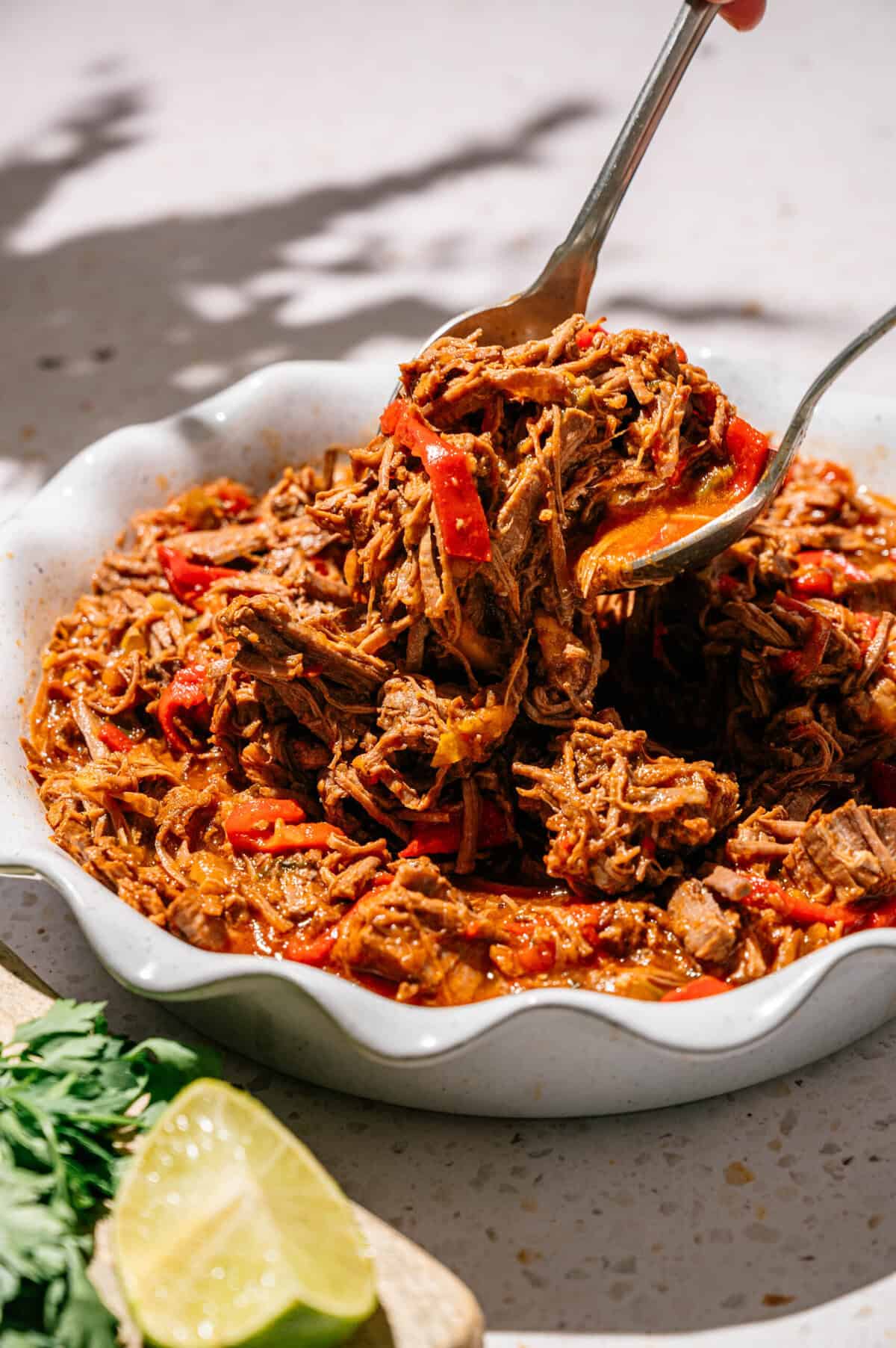A bowl of shredded beef with red peppers in a rich sauce, being served with tongs. Fresh cilantro and a lime wedge sit nearby on the table.