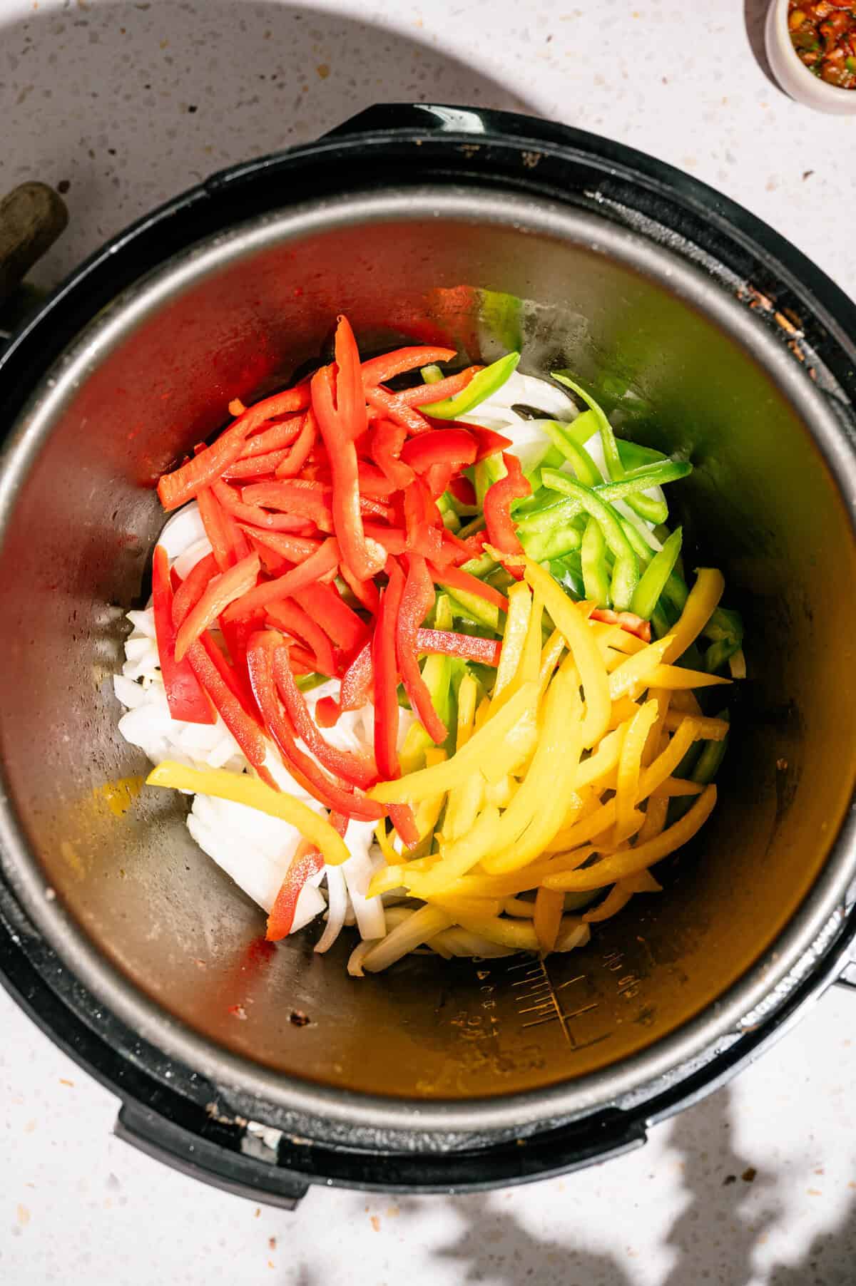 Sliced red, green, and yellow bell peppers with onions inside an Instant Pot, ready to be cooked. Small bowl with seasonings is visible in the background.