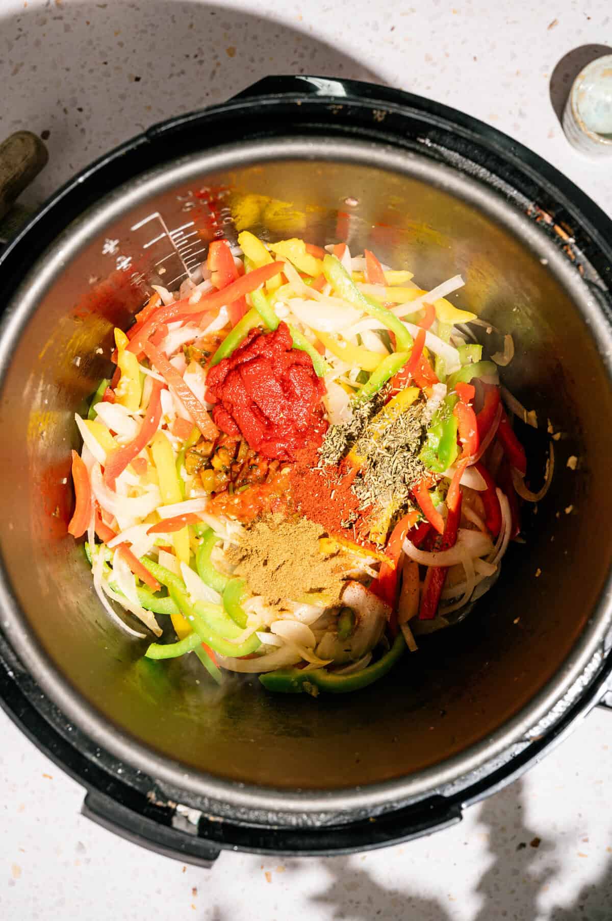 Sliced onions, green and red bell peppers, tomato paste, and spices are arranged in an Instant Pot on a white countertop, ready to be cooked.