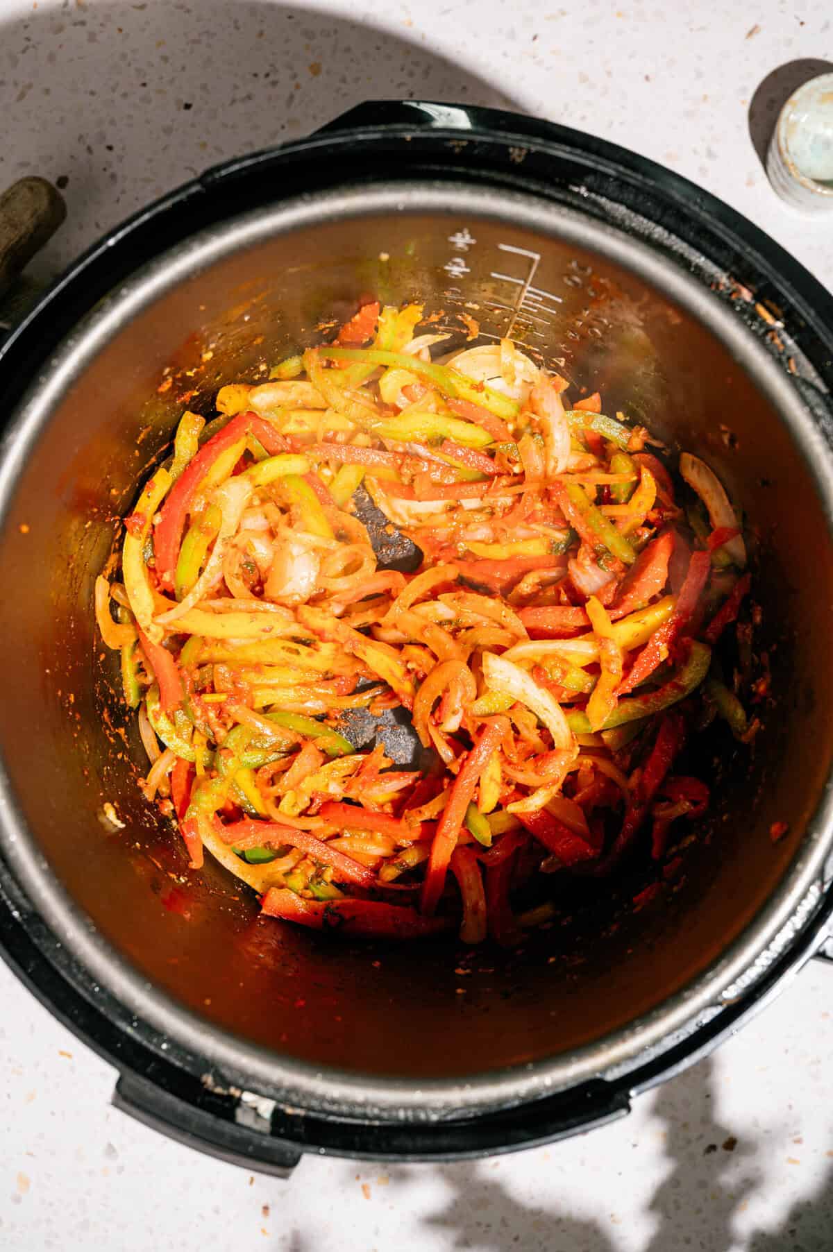 Sautéed sliced bell peppers and onions in an Instant Pot, with seasonings visible, on a speckled countertop under bright lighting.