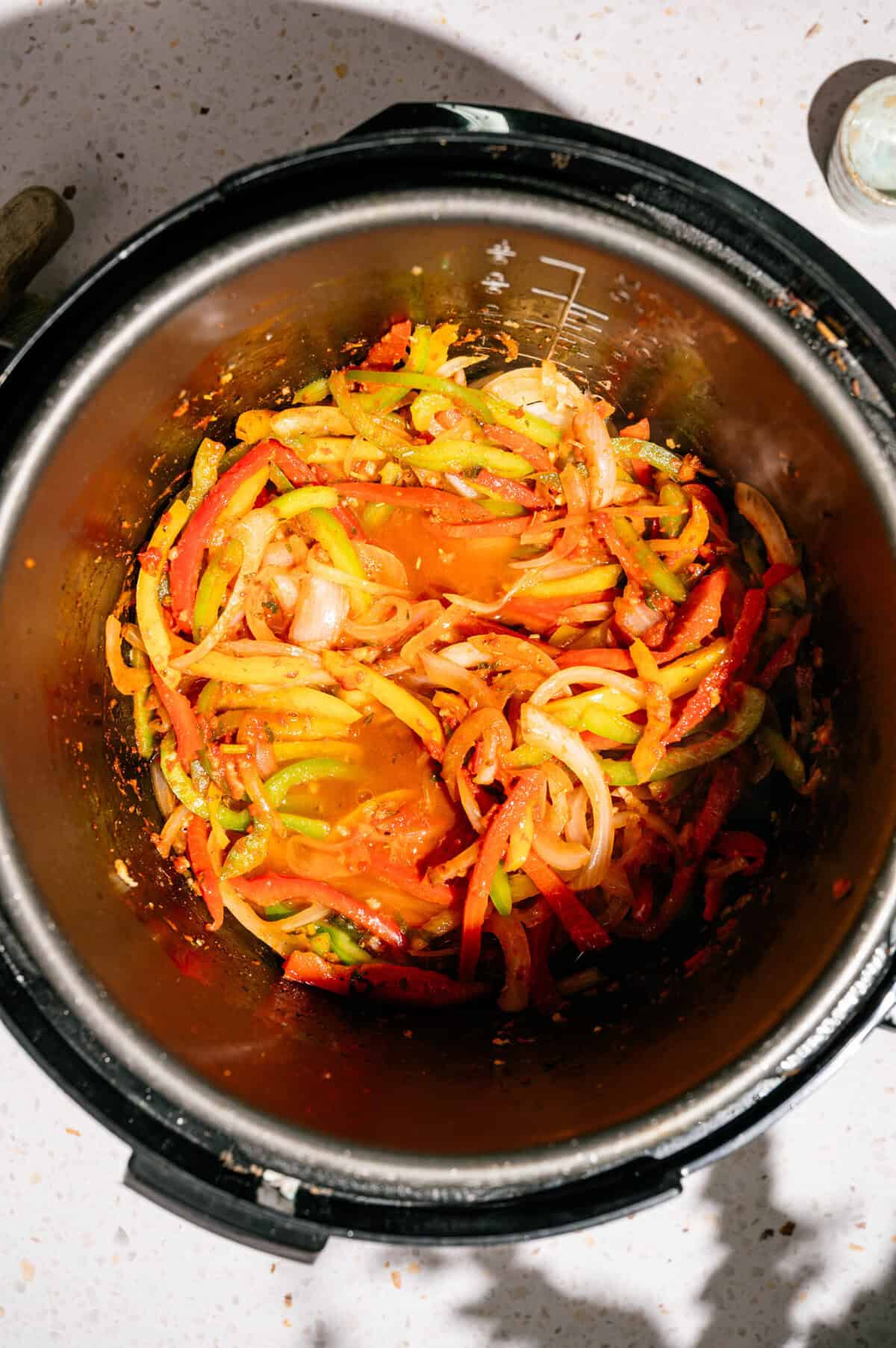 Sautéed sliced bell peppers and onions in an Instant Pot, with seasonings visible, on a speckled countertop under bright lighting.