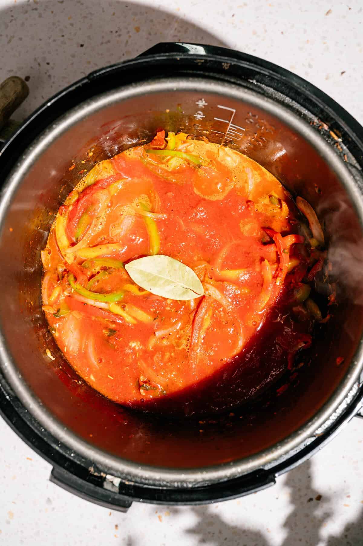 Tomato-based stew with onions and a bay leaf on top, cooking inside an Instant Pot or electric pressure cooker, viewed from above on a light countertop.