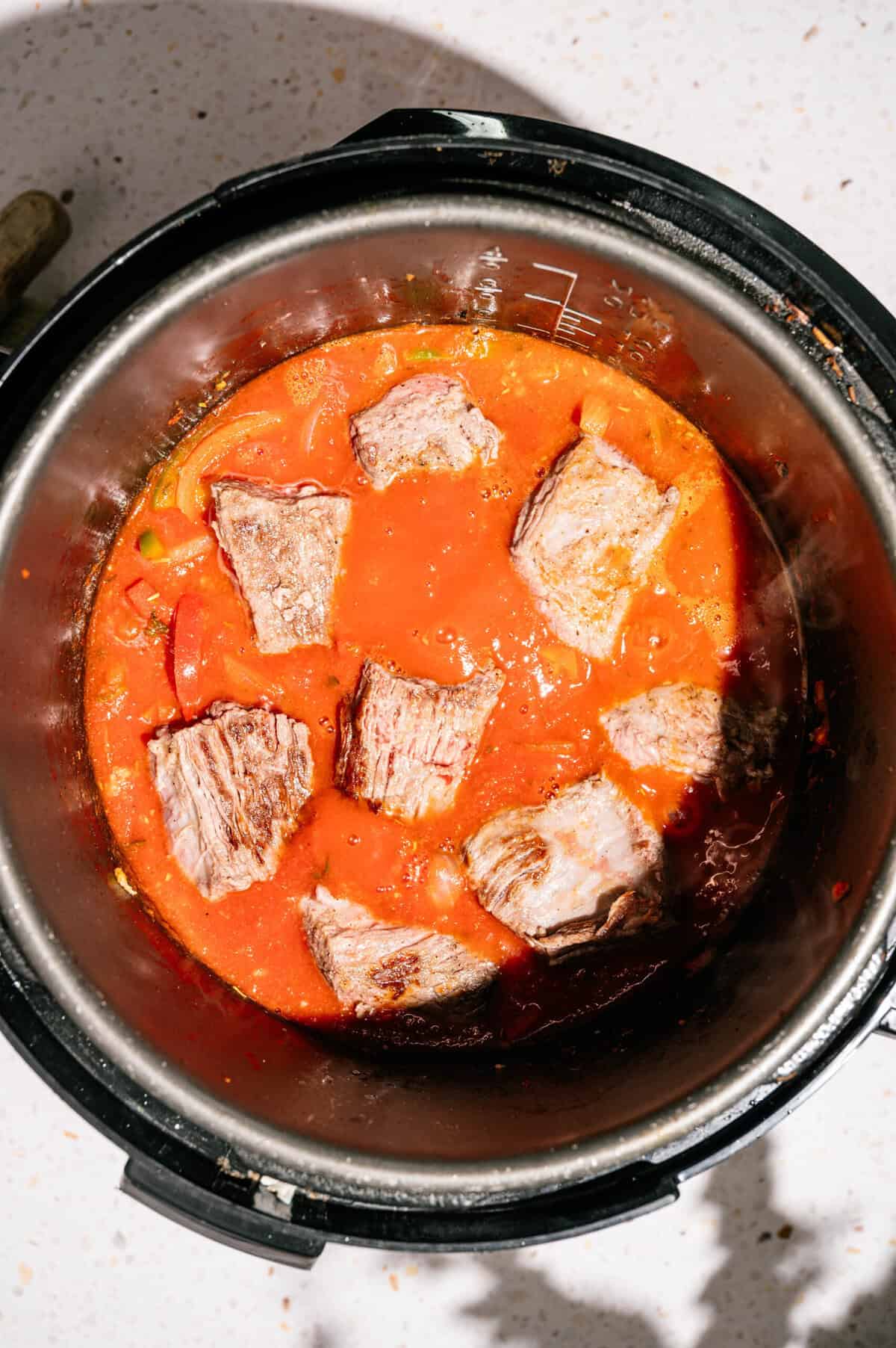 Chunks of beef simmer in a rich red tomato sauce inside an open Instant Pot, sitting on a light-colored countertop.