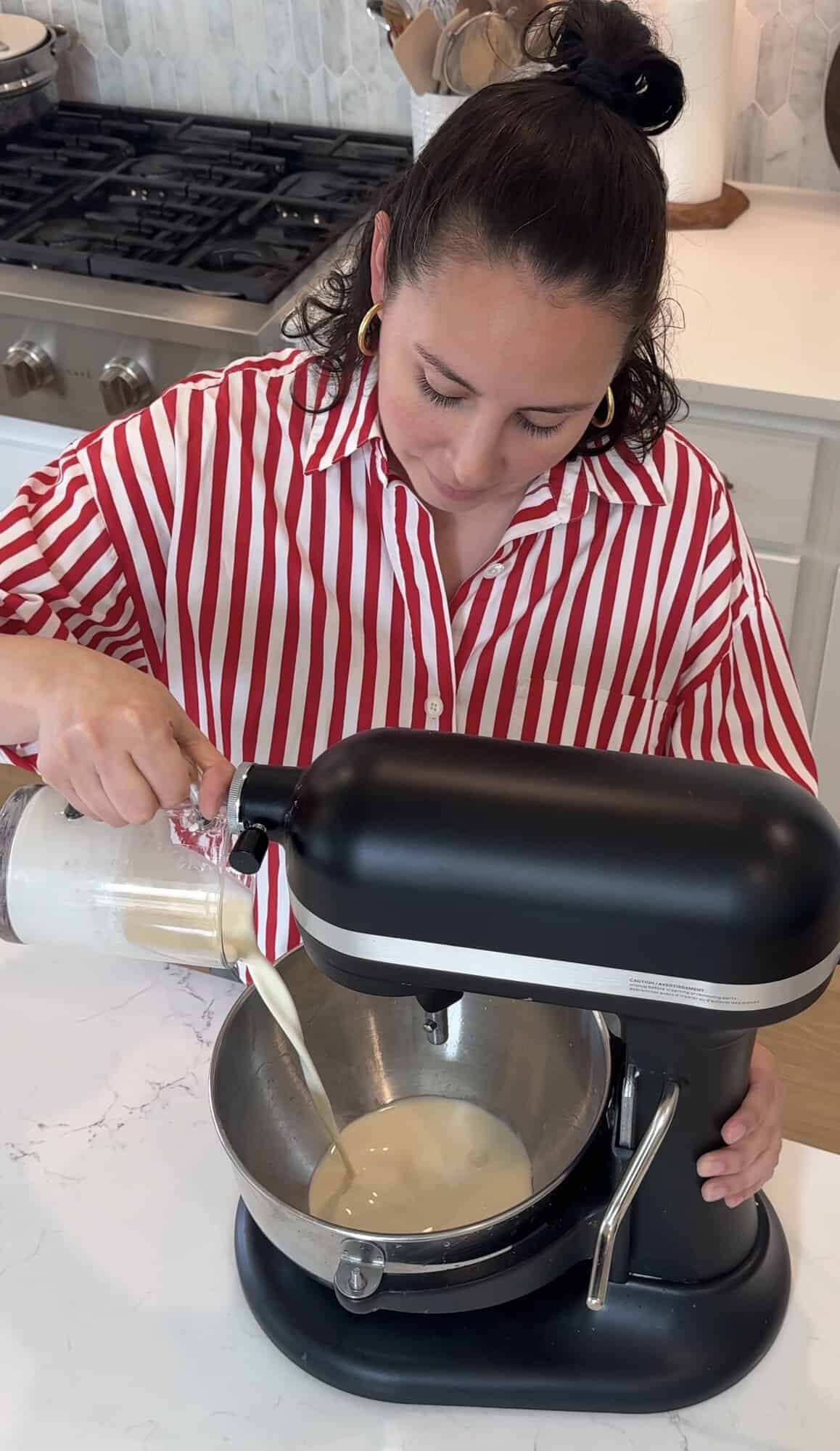 A woman in a red and white striped shirt pours liquid from a measuring cup into a stand mixer bowl in a kitchen.
