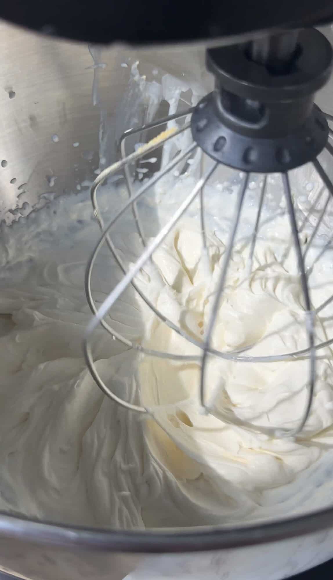 A close-up of a stand mixers wire whisk attachment beating thick, creamy whipped mixture in a metal mixing bowl.