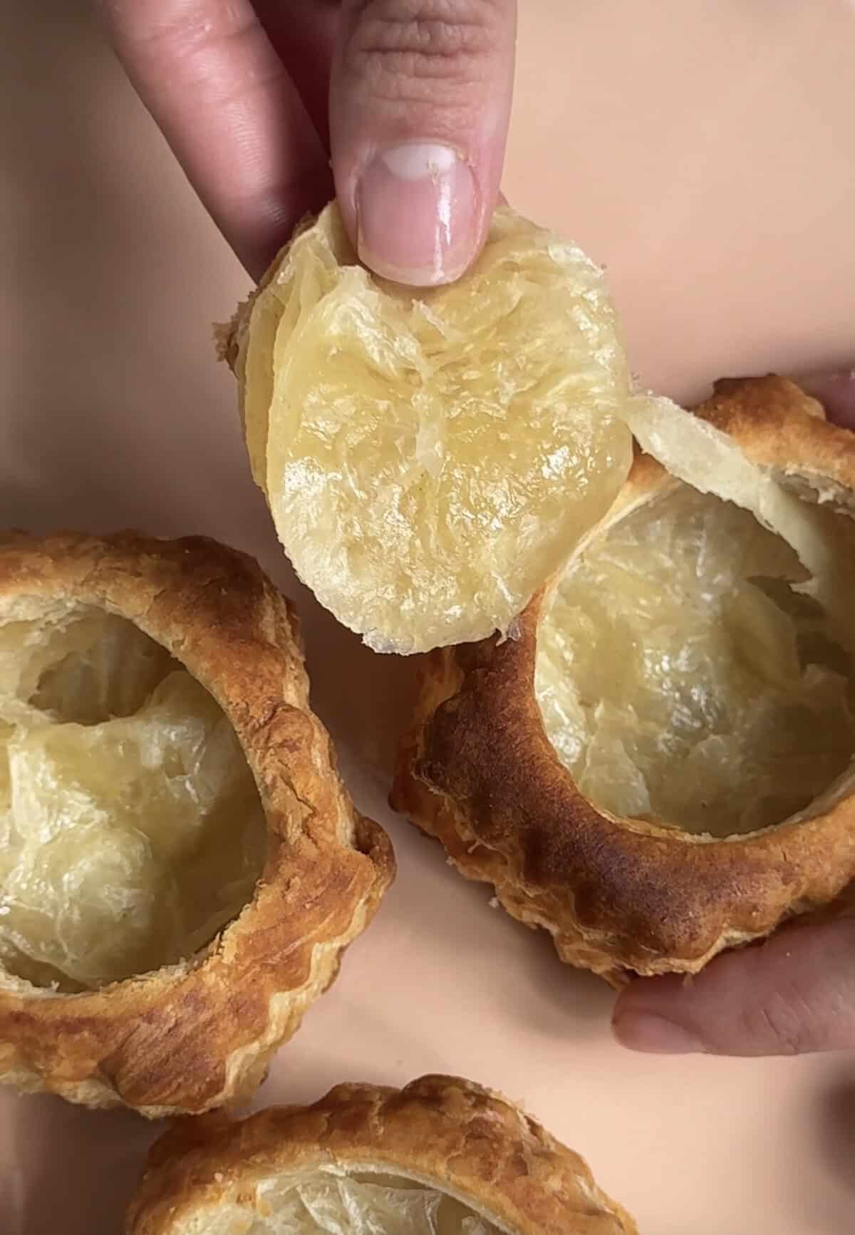 A hand lifts the flaky lid off a round, golden-brown puff pastry shell, revealing the hollow interior. Two more pastry shells are visible on a light surface.