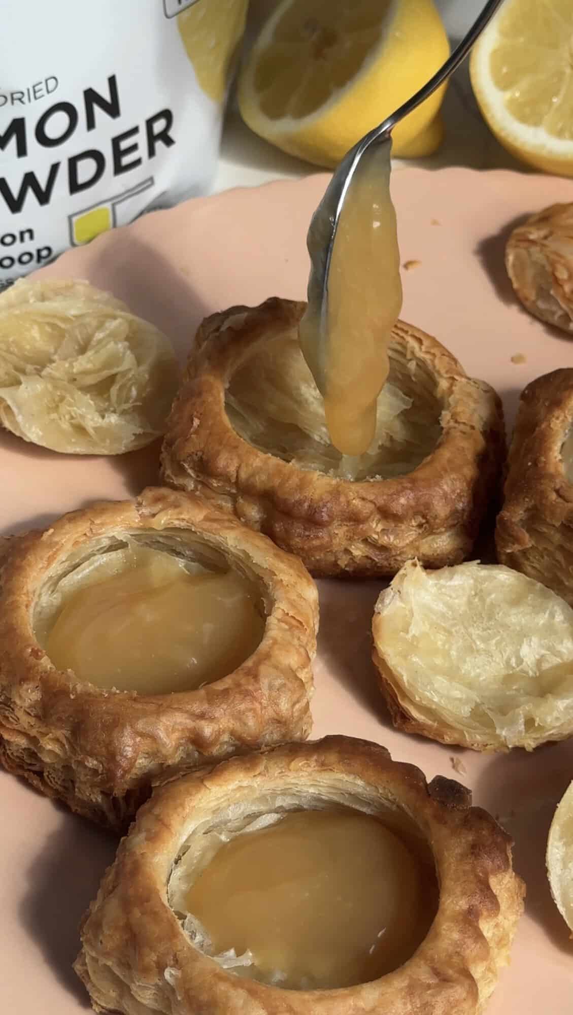 A spoon pours lemon curd into hollow puff pastry rounds, with squeezed lemon halves and a lemon powder container nearby on a peach-colored surface.