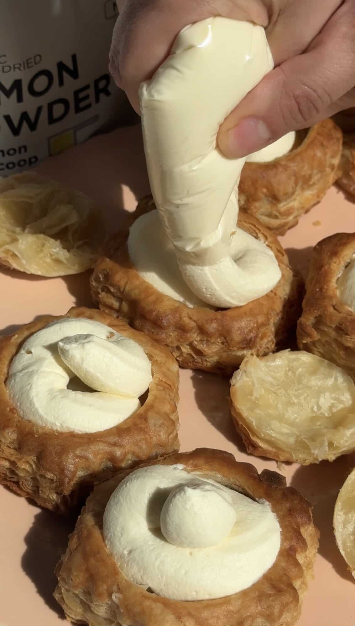 A hand pipes cream filling into golden pastry shells on a peach-colored surface, with some pastries already filled and others open, ready to be filled.