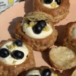 A hand placing a puff pastry shell filled with cream and topped with blueberries and lemon zest on a pink plate, surrounded by similar pastries. A lemon powder packet is partially visible in the background.